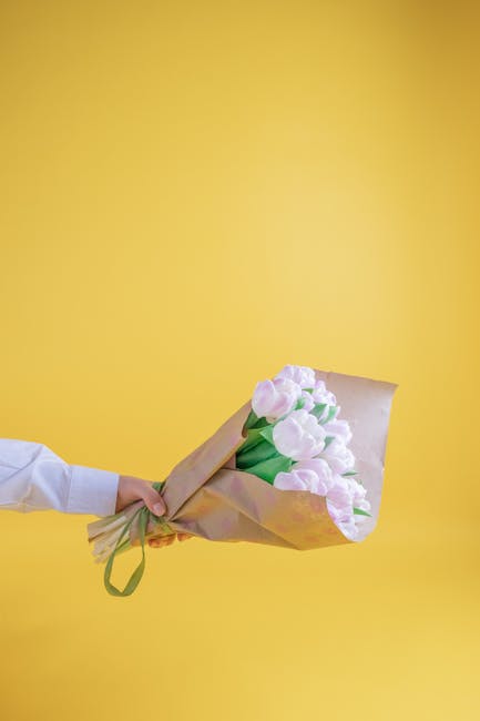 A person extends their hand holding a bouquet of fresh, pale pink tulips wrapped in light brown paper with a green ribbon against a bright yellow background. The tulips have smooth, rounded petals and