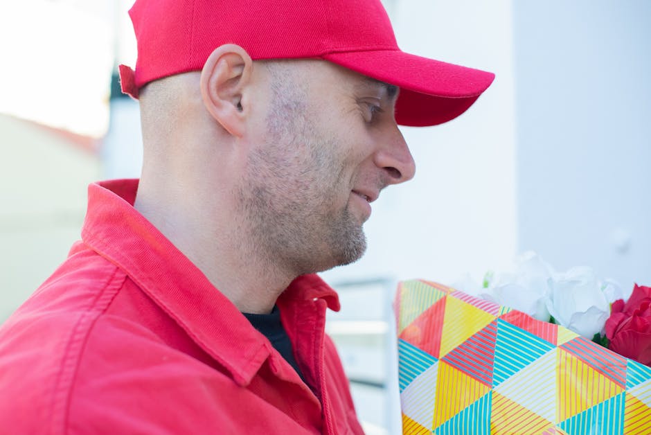 A delivery person in a red uniform and matching cap smiling as he holds a colorful floral gift box with a geometric pattern, filled with white and pink flowers, during a flower delivery service, with 