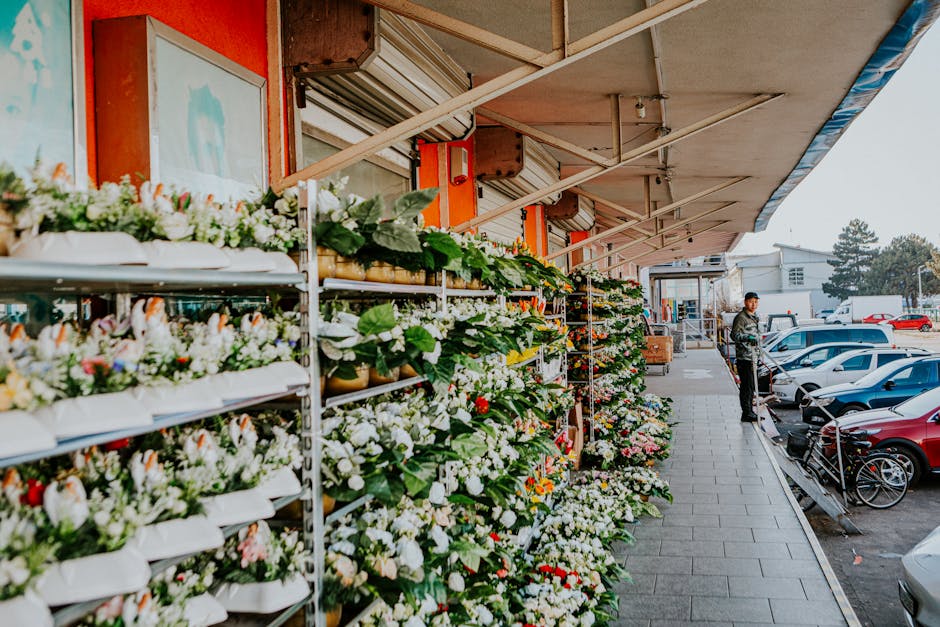 A floral display outside a flower shop, showcasing an extensive arrangement of fresh flowers including white roses, pink tulips, and mixed blossoms arranged on tiered metal shelves. The flowers are wr