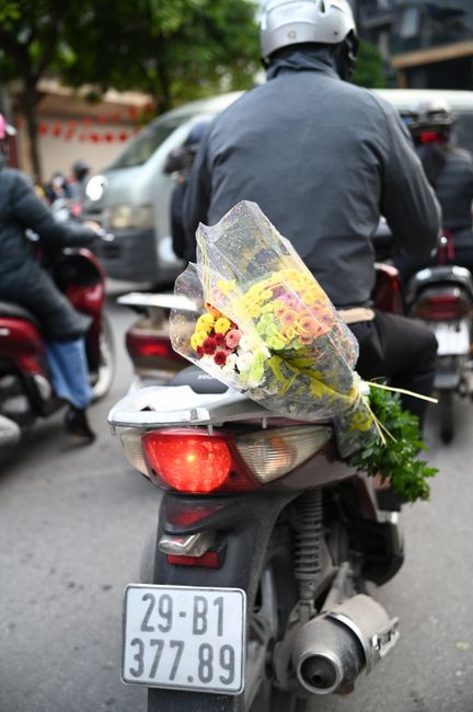 A delivery person wearing a grey helmet and jacket is seated on a black scooter, holding a bouquet of fresh flowers secured in transparent wrapping. The bouquet includes yellow and red flowers, comple