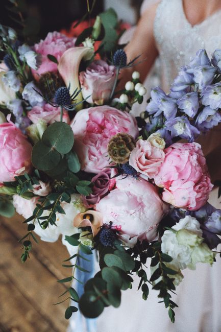 A close-up of a bridal floral bouquet featuring large, soft pink peonies with ruffled petals, interspersed with smaller lavender roses and delicate white blooms. The arrangement includes dark blue thi
