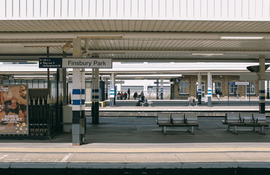Empty Finsbury Park train station platform with a white canopy overhead, featuring blue and white striped signage. Several wooden and metal benches are positioned along the platform, and a few passeng